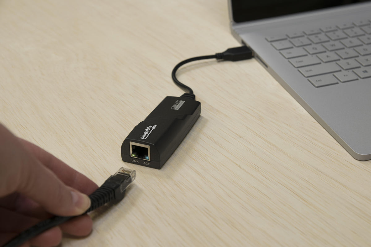 Plugable USB 2.0 Gigabit LAN adapter connected to a laptop, showing an Ethernet port with LINK and ACT status lights.